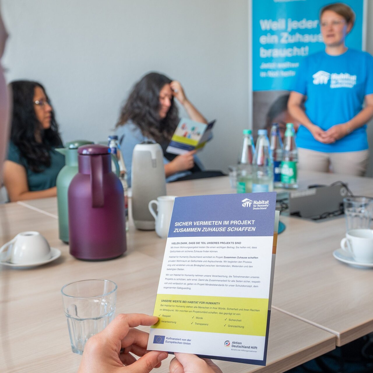 Im Besprechungsraum hält eine Person das Faltblatt „Sicher vermieten im Projekt Zusammen Zuhause schaffen" in den Händen.