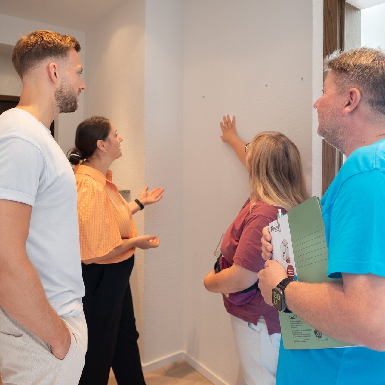 Four people stand in the hallway of an empty apartment; one of them points at the wall.