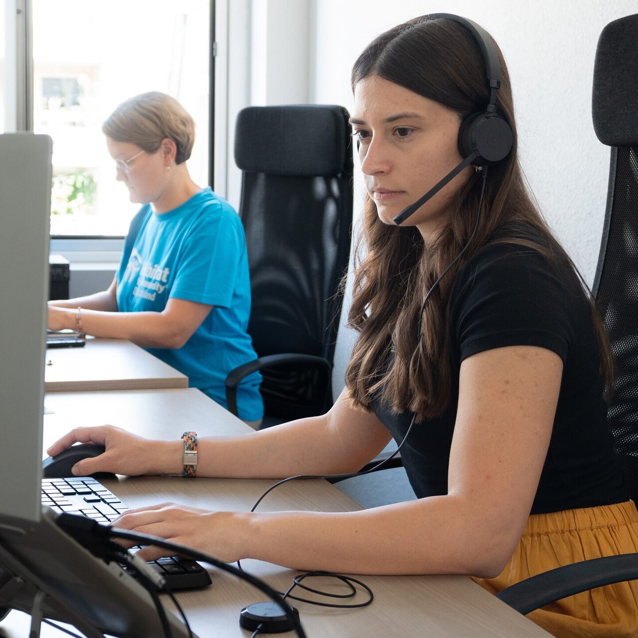A staff member with a headset works at a computer; in the background a colleague in a Habitat T-shirt.