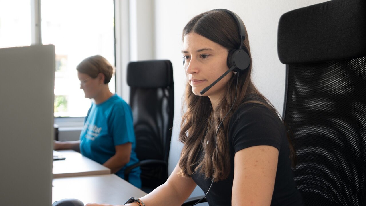 A staff member with a headset works at a computer; in the background a colleague in a Habitat T-shirt